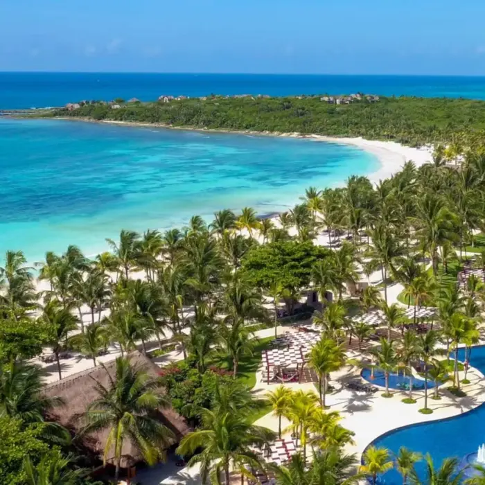 aerial view of Barceló Maya Beach resort, showcasing large pools, lush palm trees, and the expansive white sand beach bordering the turquoise ocean.