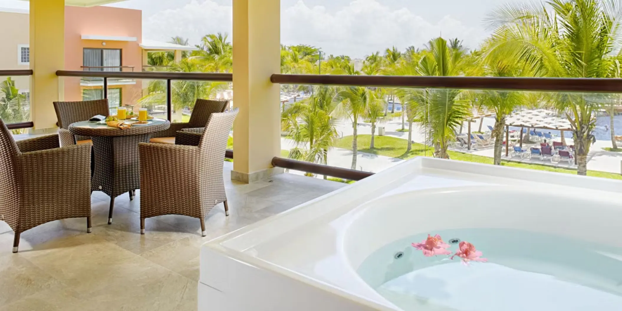 A suite balcony featuring a hot tub and a comfortable dining area with wicker chairs, overlooking the resort's lush grounds and pools.