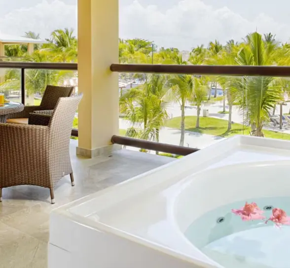 A suite balcony featuring a hot tub and a comfortable dining area with wicker chairs, overlooking the resort's lush grounds and pools.