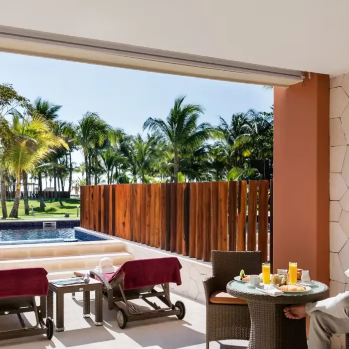 couple relaxing on the patio of a swim-out suite; one reading at a dining table, the other standing by the private pool overlooking the resort's grounds.