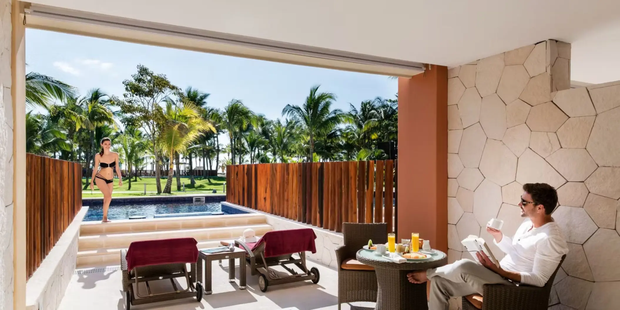 couple relaxing on the patio of a swim-out suite; one reading at a dining table, the other standing by the private pool overlooking the resort's grounds.