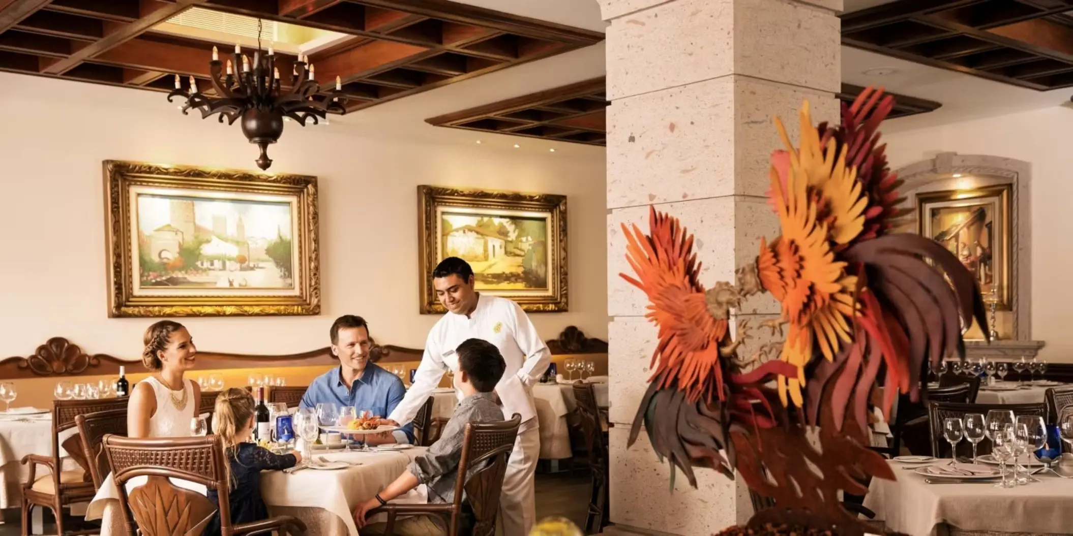 A family enjoying a meal at an upscale restaurant, with a server attending to them in a room with ornate chandeliers and elegant decor.