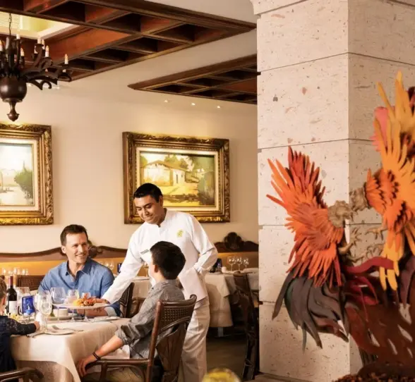 A family enjoying a meal at an upscale restaurant, with a server attending to them in a room with ornate chandeliers and elegant decor.