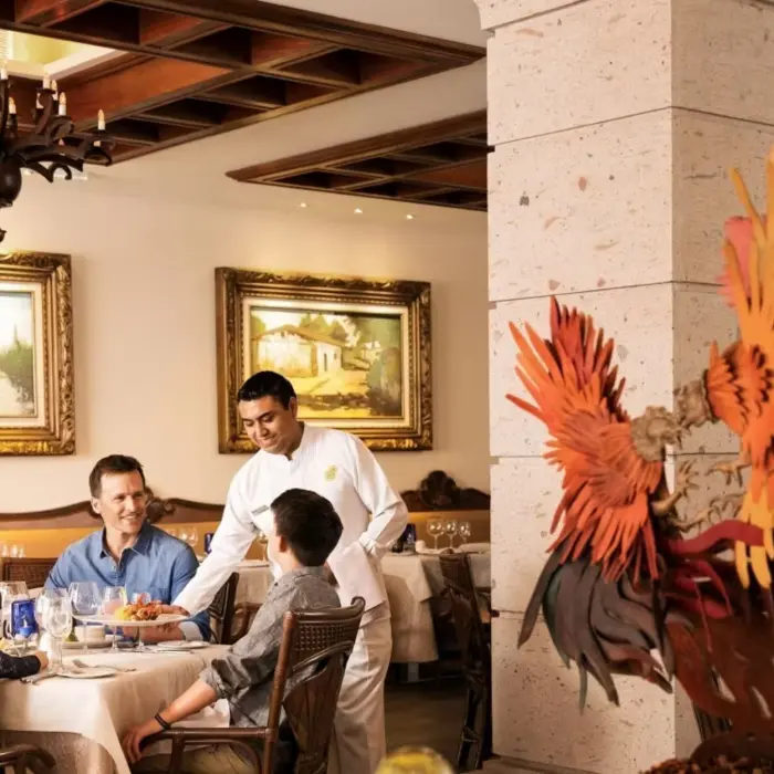 A family enjoying a meal at an upscale restaurant, with a server attending to them in a room with ornate chandeliers and elegant decor.