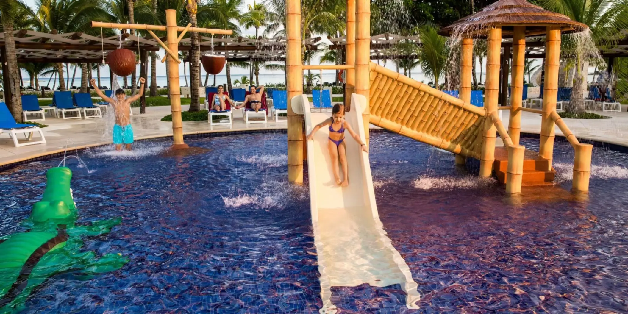 Children enjoying a colorful water park area with a bamboo slide, water features, and a large crocodile sculpture in a shallow pool designed for kids.
