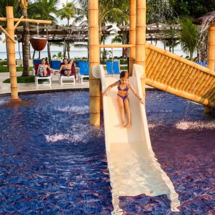 Children enjoying a colorful water park area with a bamboo slide, water features, and a large crocodile sculpture in a shallow pool designed for kids.