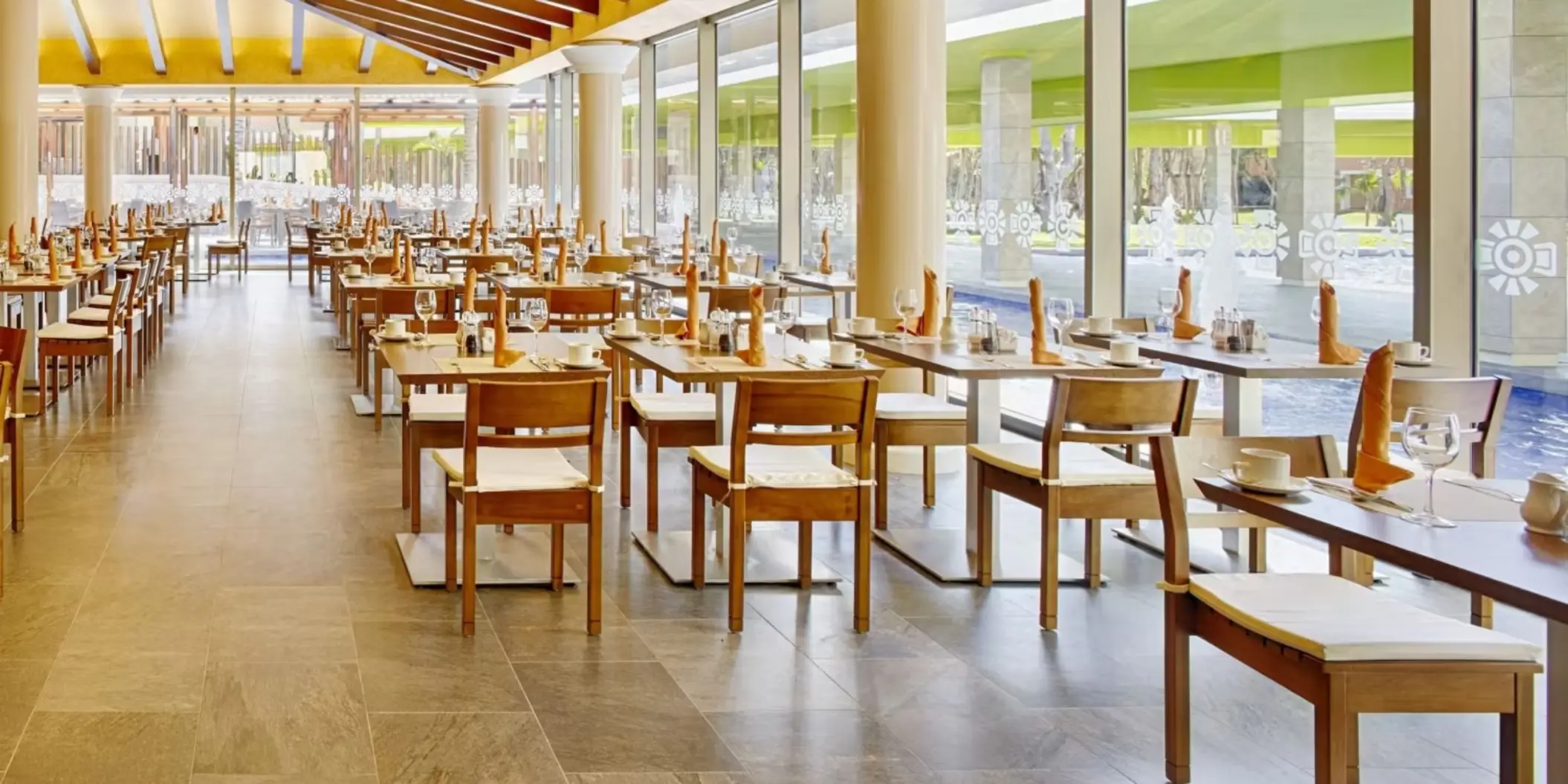 A spacious indoor dining area with many tables and chairs, large windows overlooking a pool, and a view of the resort's outdoor area.