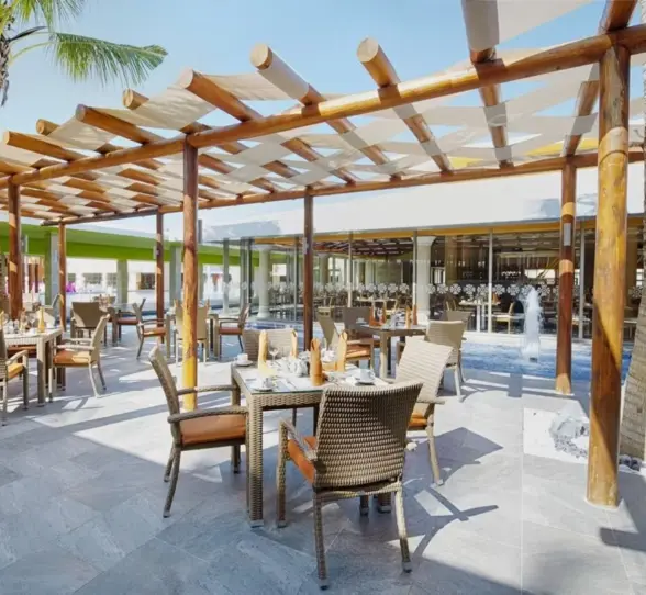 A spacious outdoor terrace dining area with wooden pergolas, comfortable seating, and tables set for dining, surrounded by palm trees and resort views.
