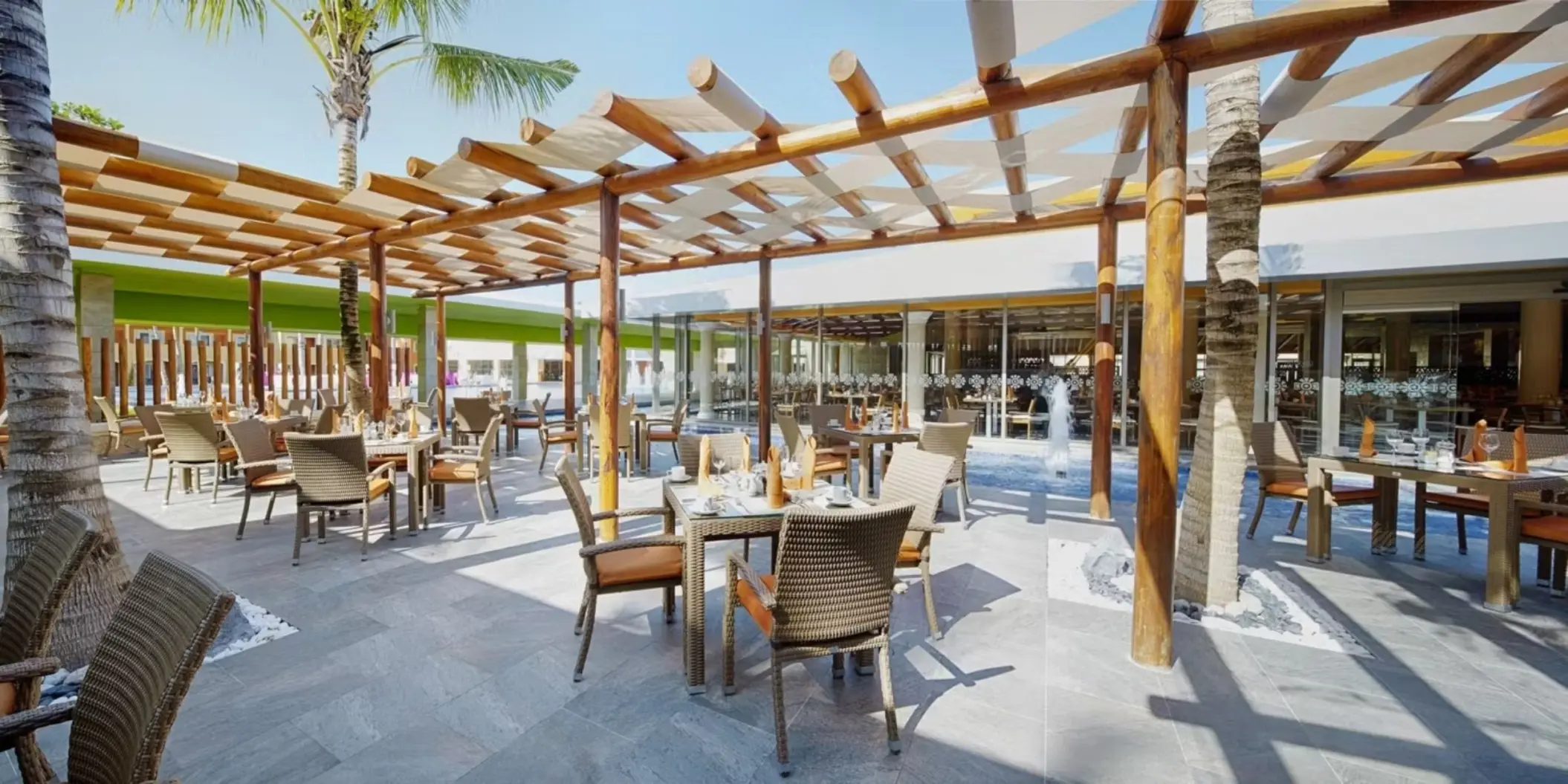 A spacious outdoor terrace dining area with wooden pergolas, comfortable seating, and tables set for dining, surrounded by palm trees and resort views.