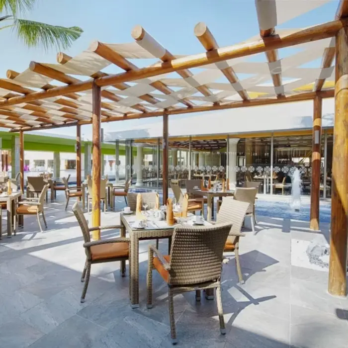 A spacious outdoor terrace dining area with wooden pergolas, comfortable seating, and tables set for dining, surrounded by palm trees and resort views.