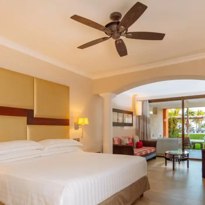 A spacious suite at Barceló Maya Beach resort, featuring a large bed with a tall headboard, a sitting area, and an outdoor terrace visible through an archway.