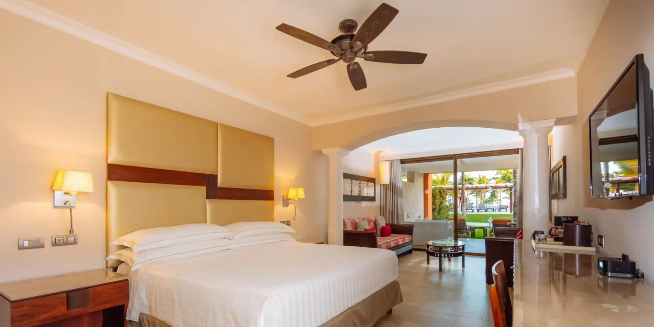 A spacious suite at Barceló Maya Beach resort, featuring a large bed with a tall headboard, a sitting area, and an outdoor terrace visible through an archway.