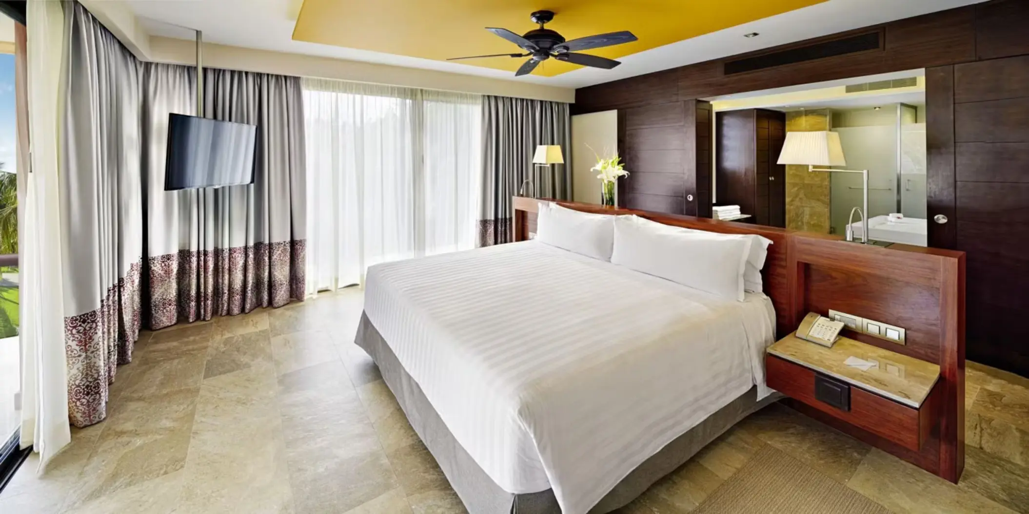modern suite with a king-size bed, dark wood accents, contemporary lighting, a mounted television, and large windows leading to a balcony.