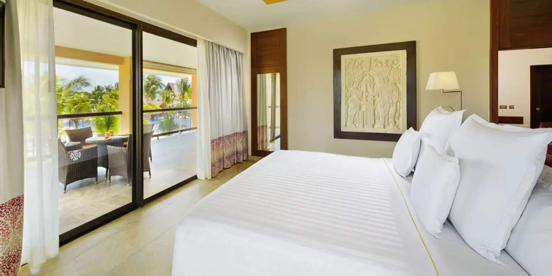 bright suite featuring a king-size bed, minimalist decor, and sliding glass doors opening onto a terrace with seating and views of the resort's exterior.