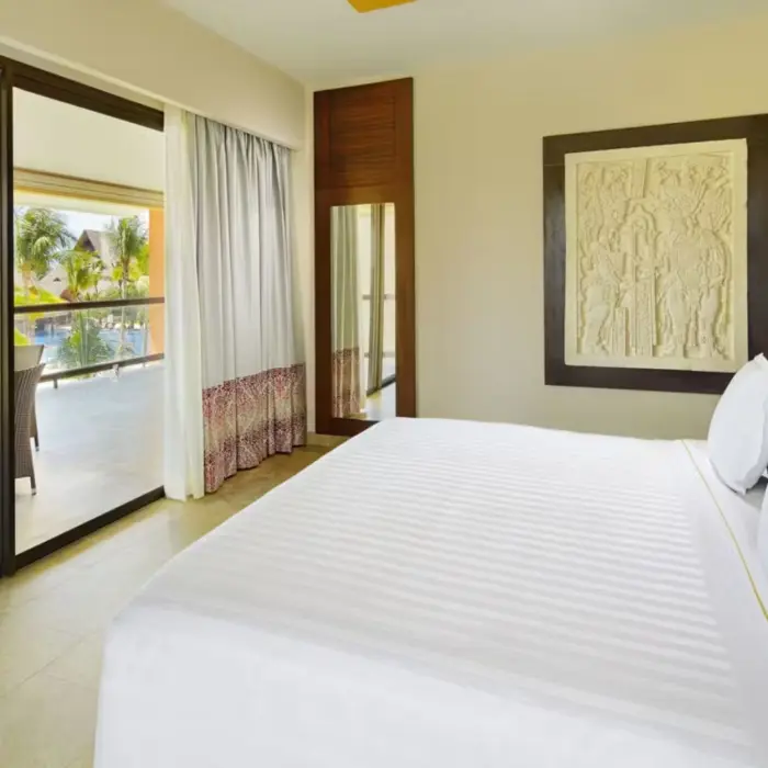 bright suite featuring a king-size bed, minimalist decor, and sliding glass doors opening onto a terrace with seating and views of the resort's exterior.