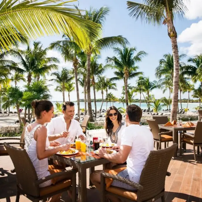 Friends dining outdoors at an oceanfront restaurant, with palm trees and the beach in the background.