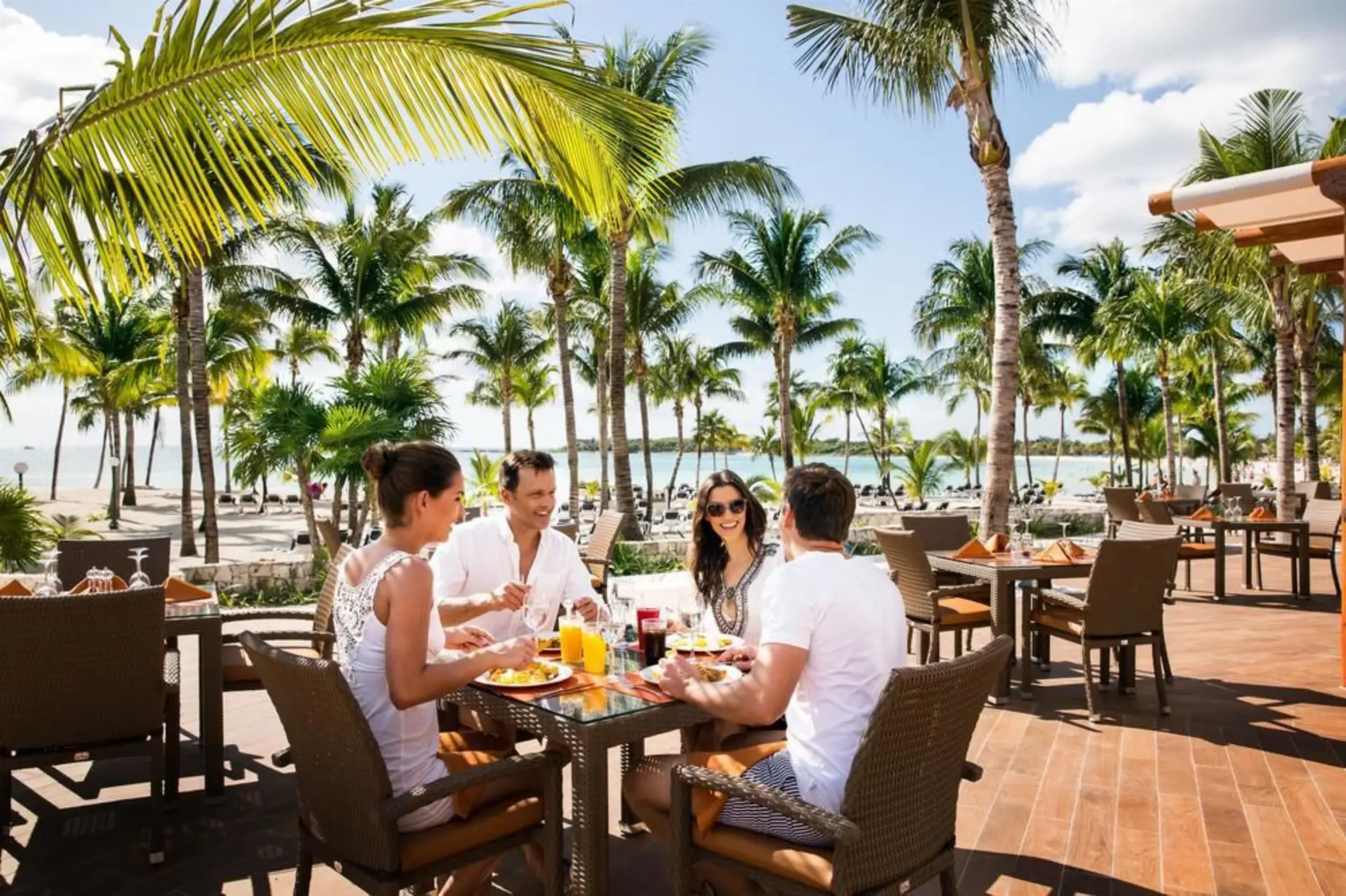 Friends dining outdoors at an oceanfront restaurant, with palm trees and the beach in the background.