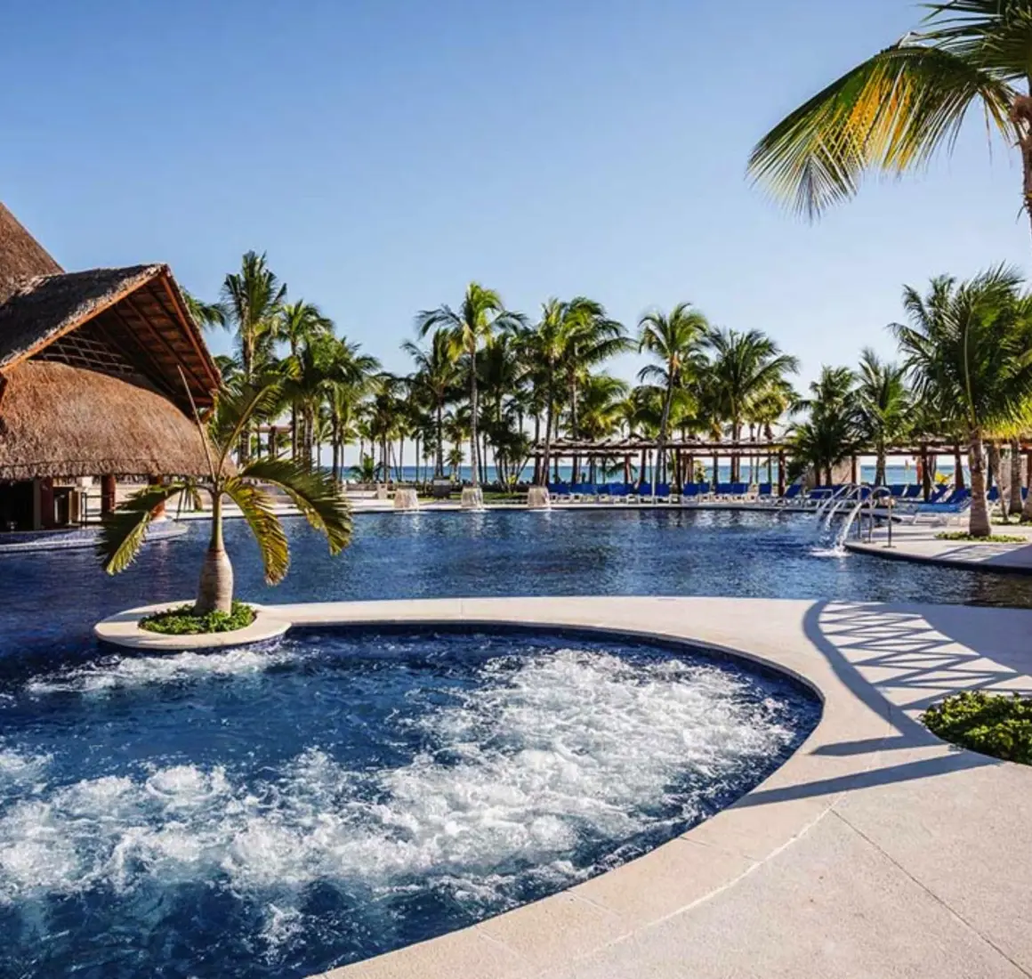 A large, inviting resort pool with a swim-up bar, surrounded by lush palm trees and cabanas.