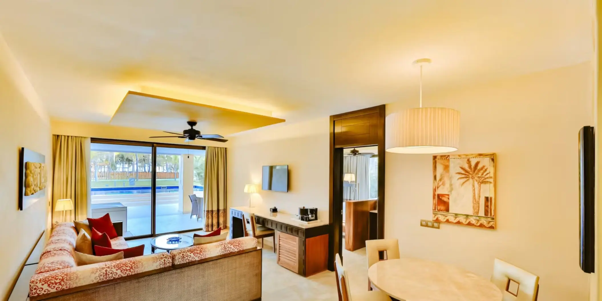 A bright and spacious hotel suite with a living area, dining table, and a balcony overlooking a pool.