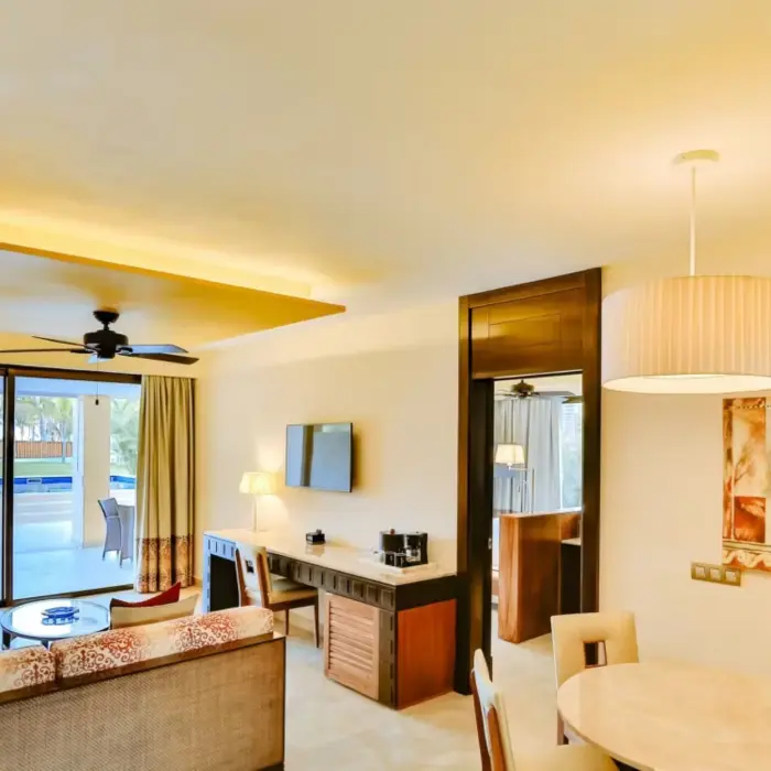 A bright and spacious hotel suite with a living area, dining table, and a balcony overlooking a pool.