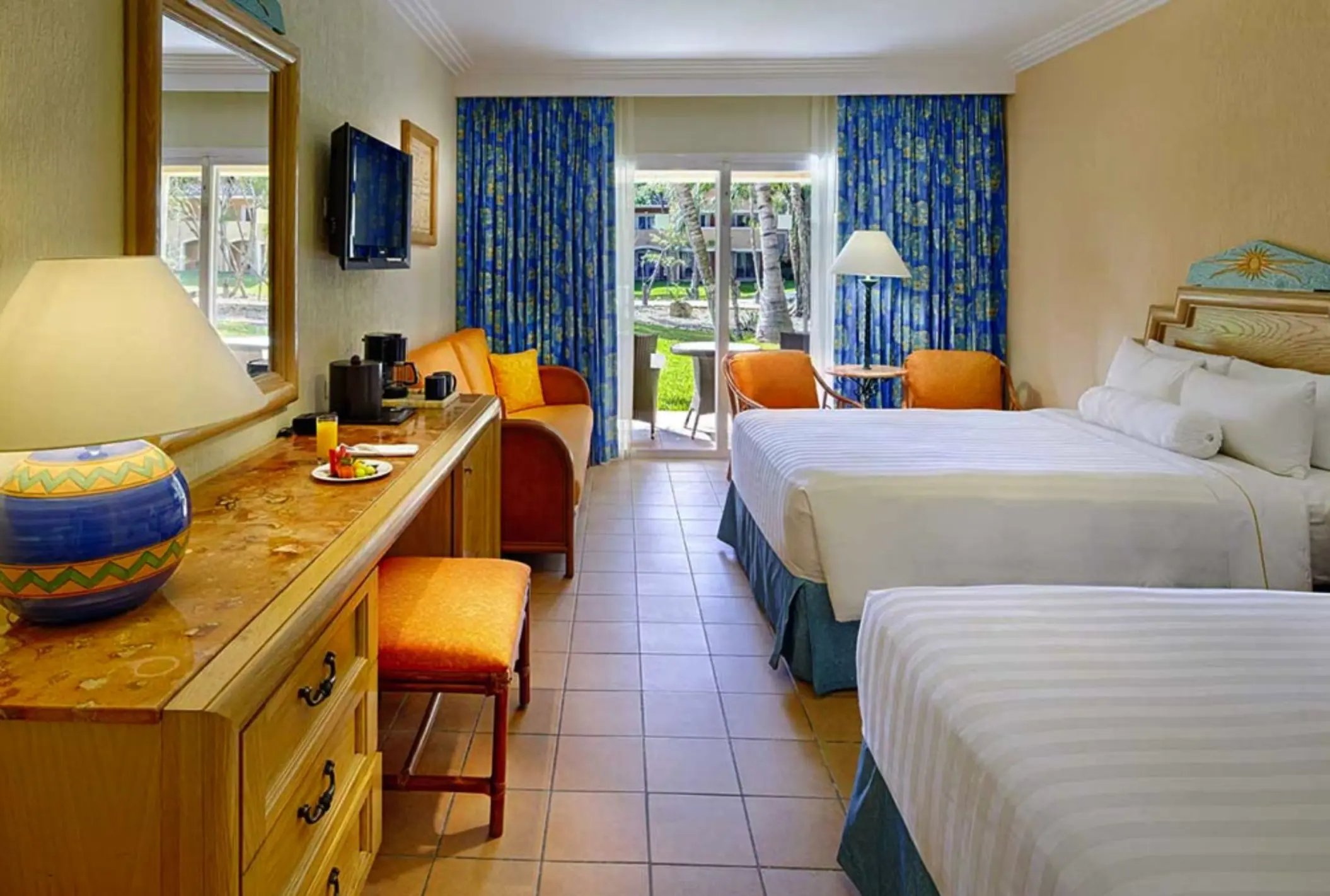 A bright hotel room with two beds, a desk area, and a balcony overlooking a garden.
