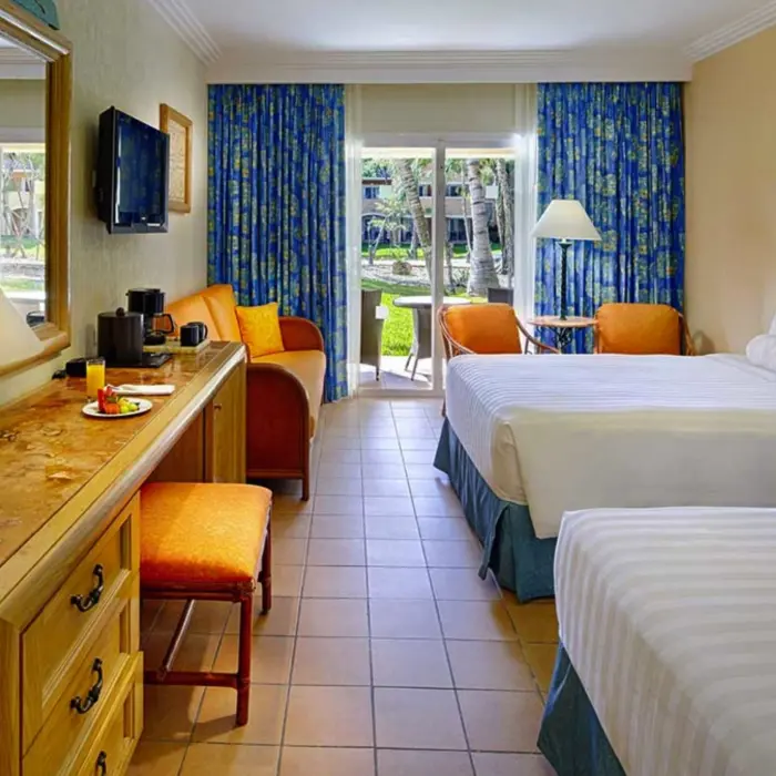 A bright hotel room with two beds, a desk area, and a balcony overlooking a garden.