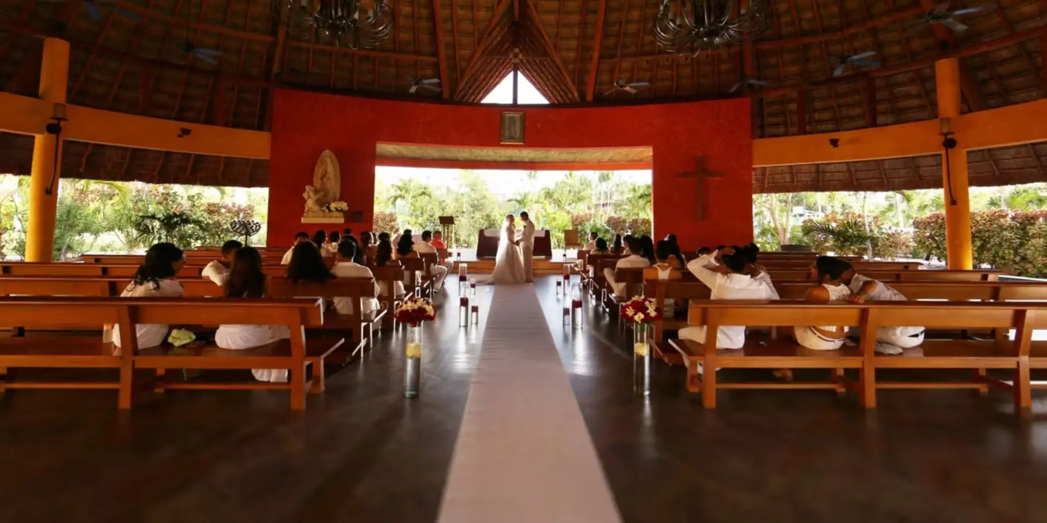 A wedding ceremony in a beautiful, open-air chapel with a wooden roof and a pathway lined with pews.
