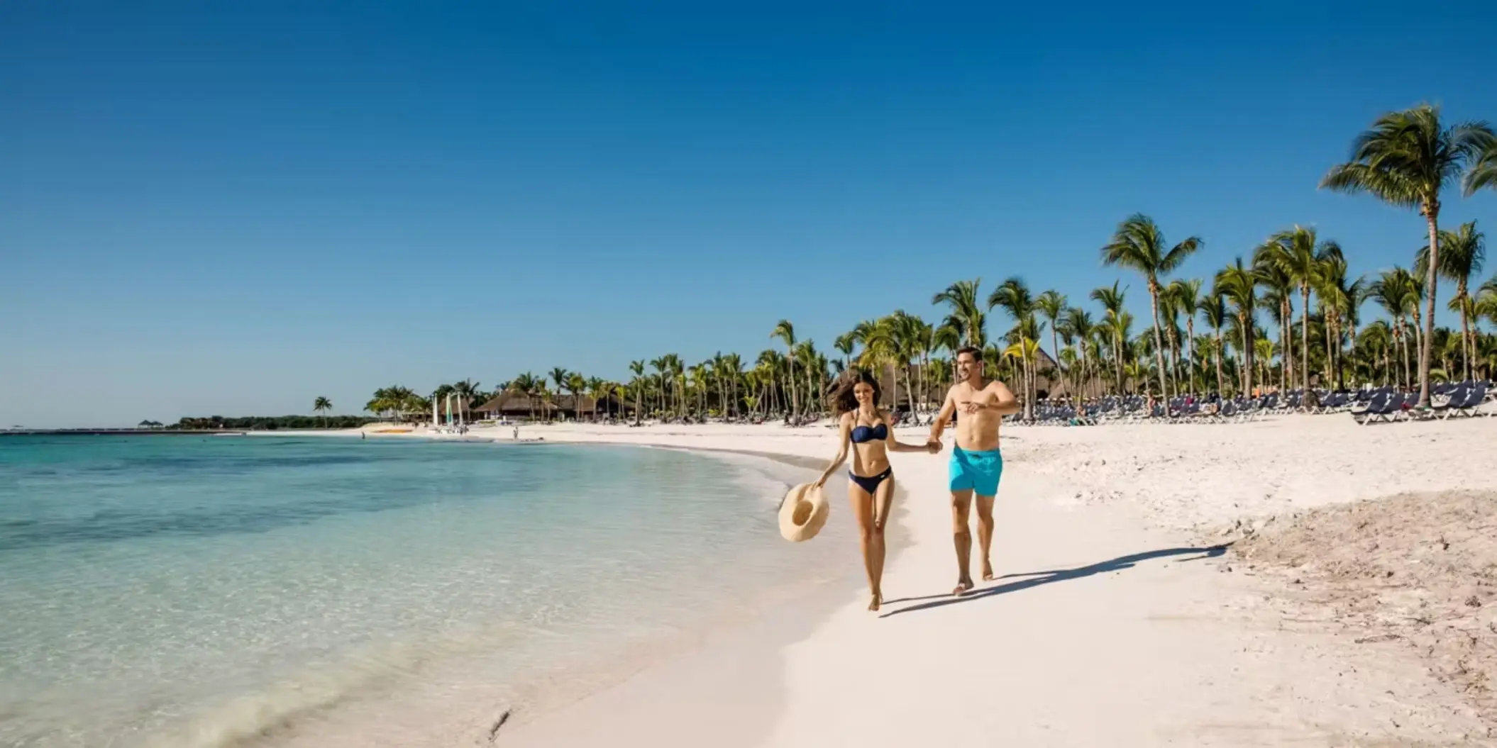 A happy couple holding hands and running along the stunning white sand beach next to the turquoise sea.