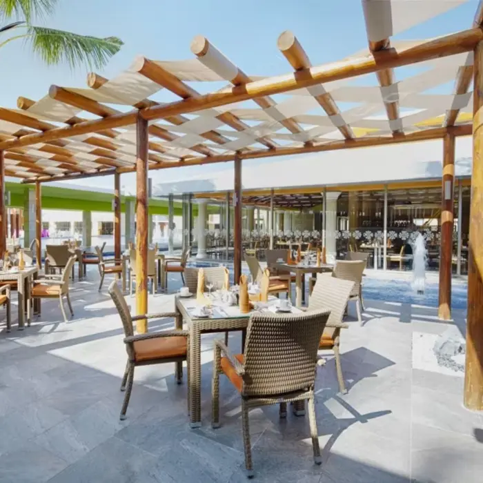 An outdoor restaurant next to a pool, with tables and chairs set under a covered wooden patio.