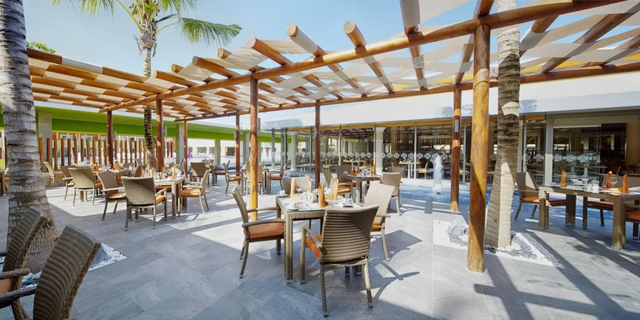 An outdoor restaurant next to a pool, with tables and chairs set under a covered wooden patio.