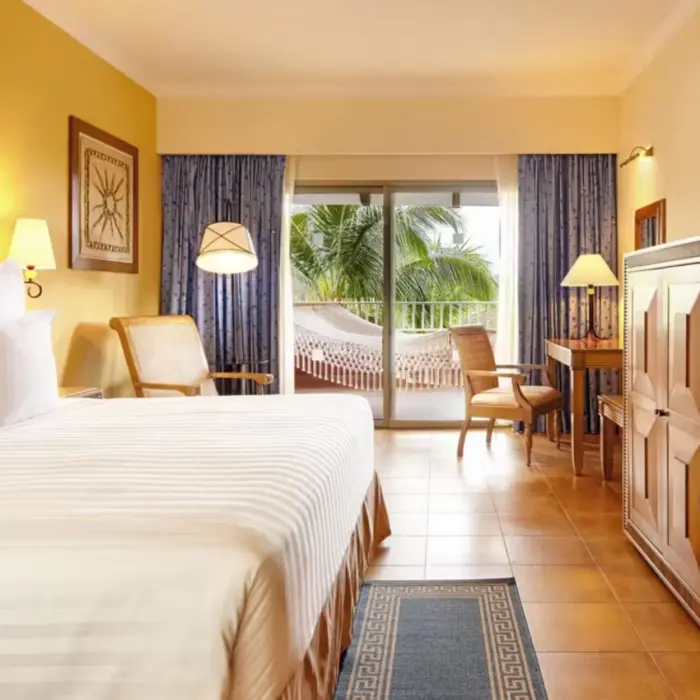 A hotel suite with a large bed and a private balcony featuring a relaxing hammock and palm tree views.