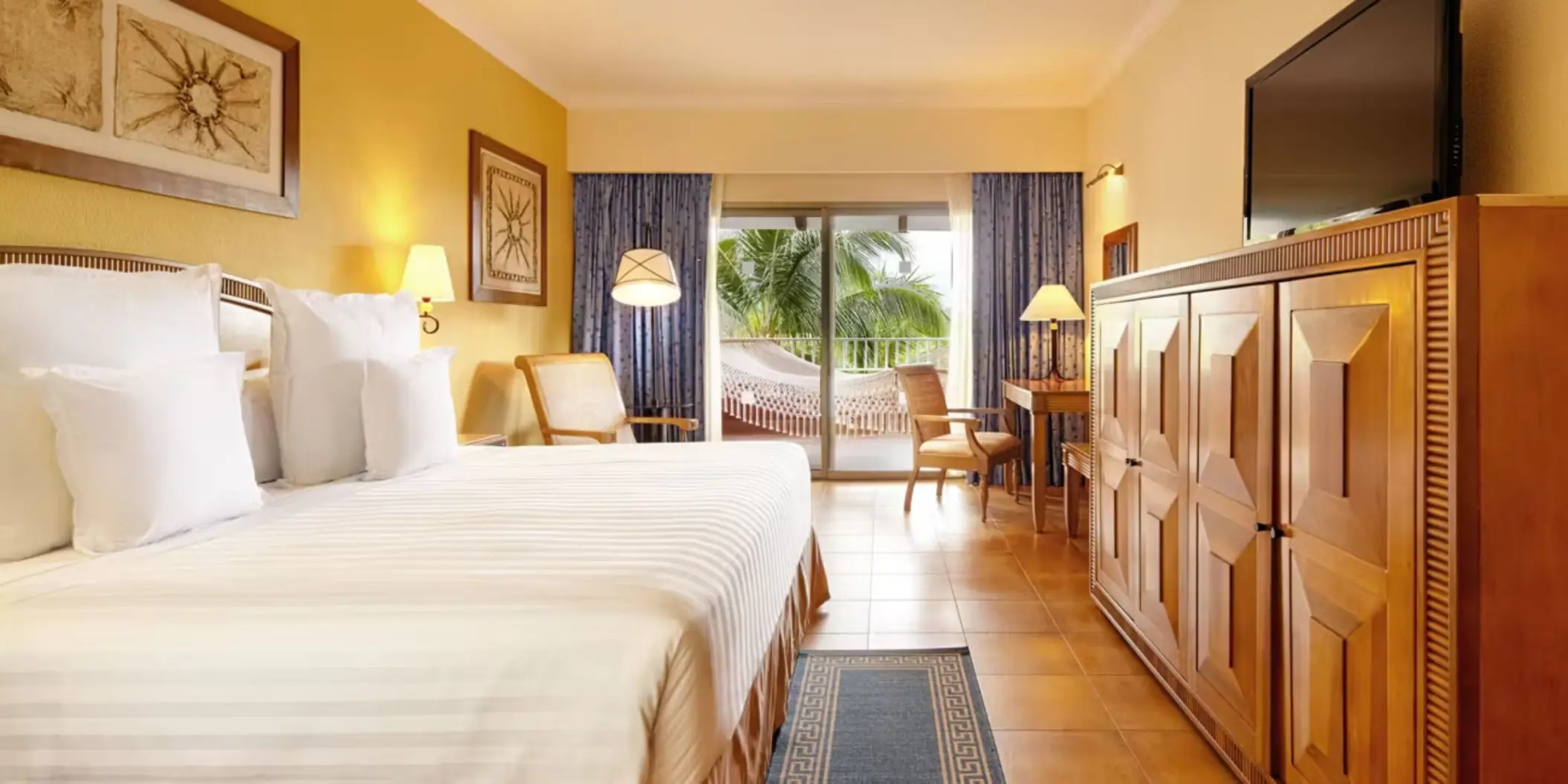A hotel suite with a large bed and a private balcony featuring a relaxing hammock and palm tree views.