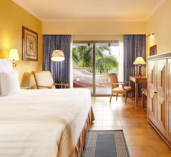 A hotel suite with a large bed and a private balcony featuring a relaxing hammock and palm tree views.
