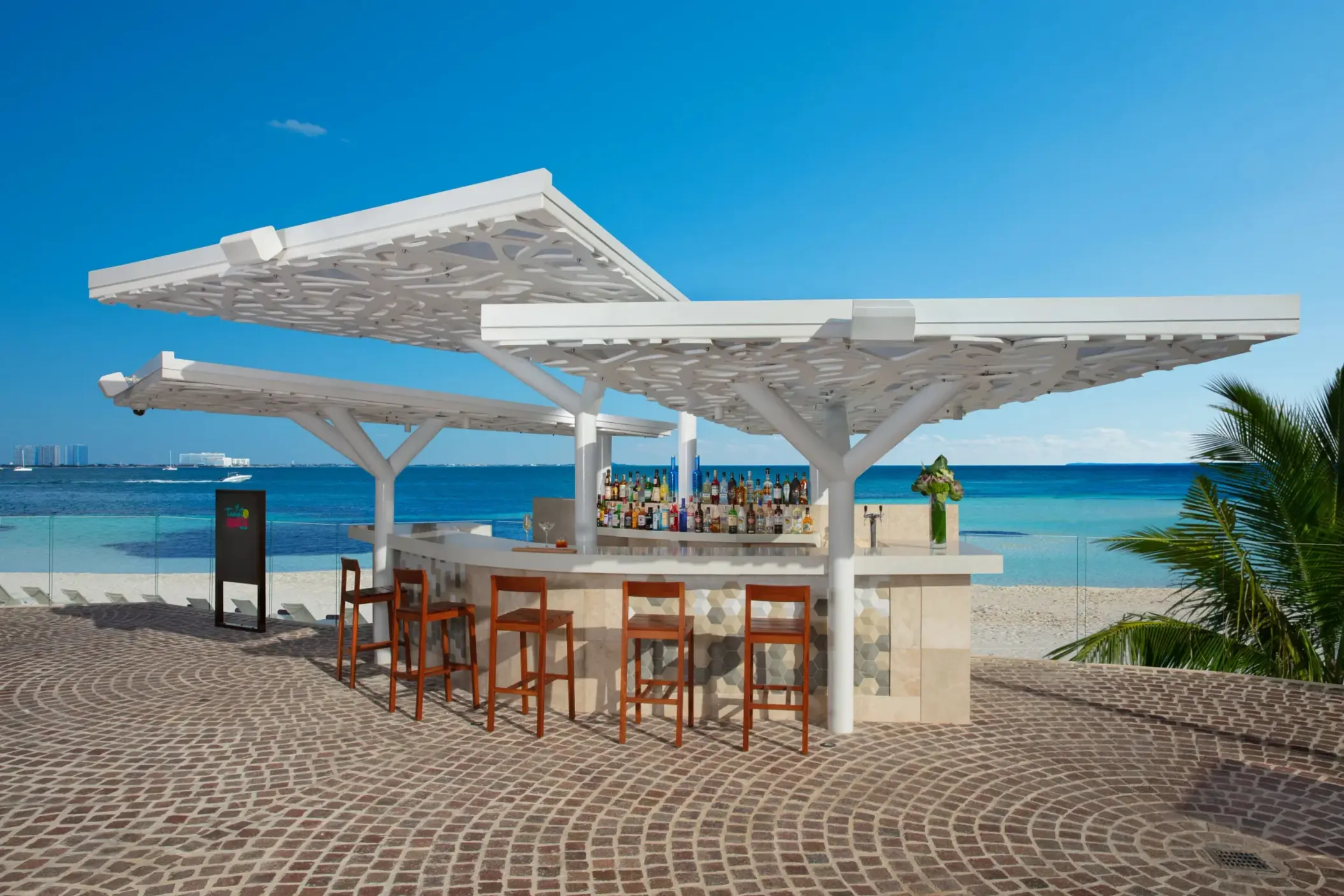 Open-air beach bar with wood stools and canopy roof, set on cobblestone patio facing turquoise water.