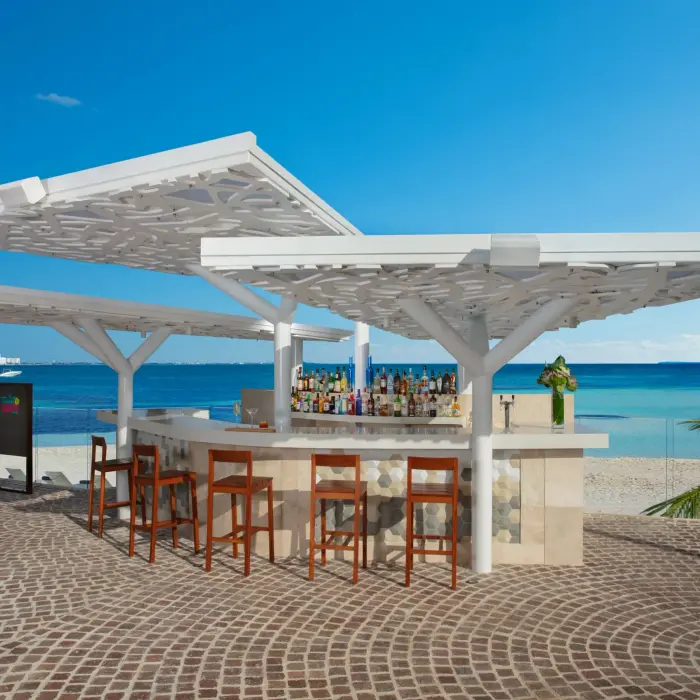 Open-air beach bar with wood stools and canopy roof, set on cobblestone patio facing turquoise water.