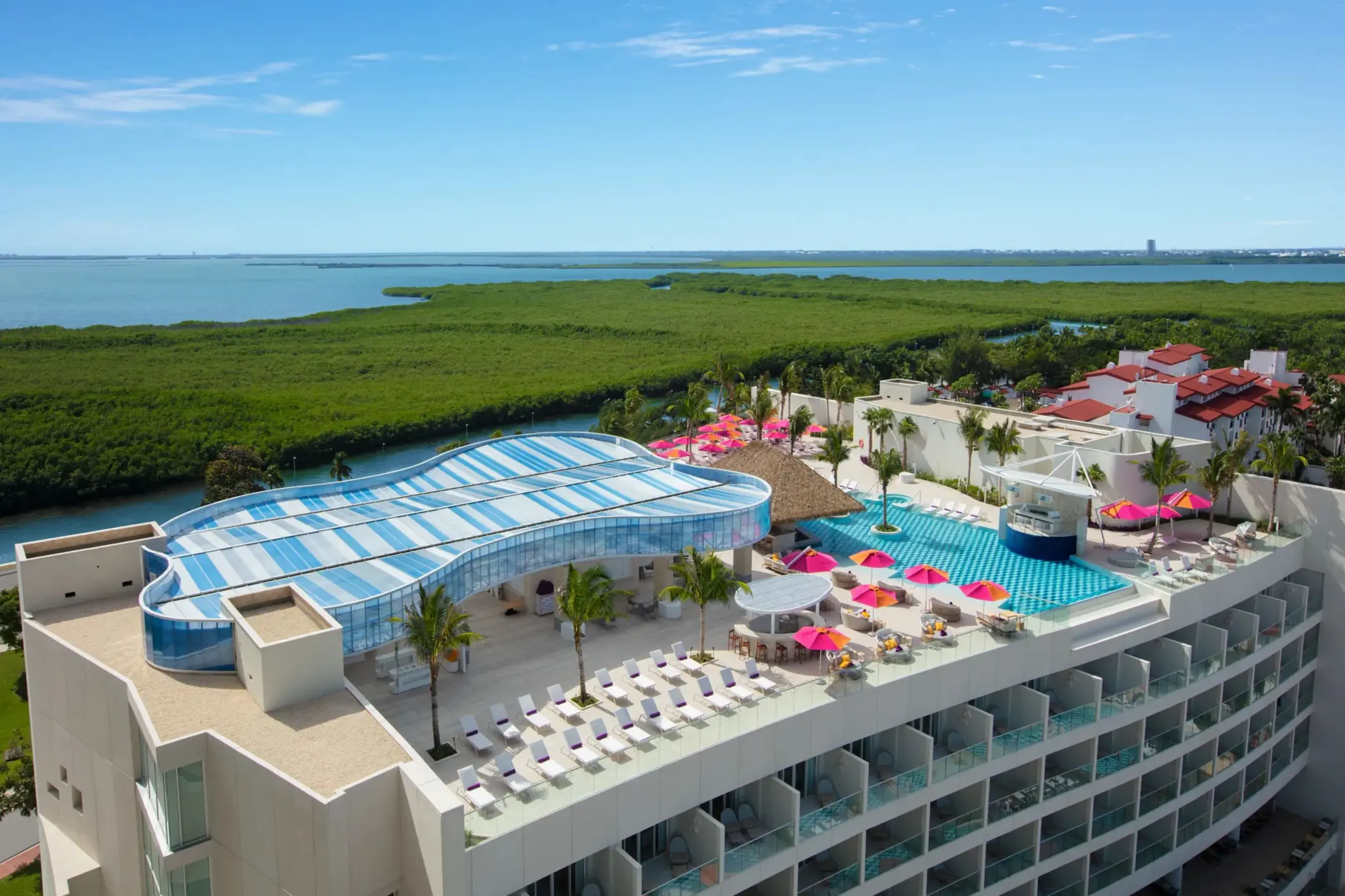 Aerial panoramic view of Xcelerate rooftop pool with lounge chairs, palm trees, and pink umbrellas by the mangroves.