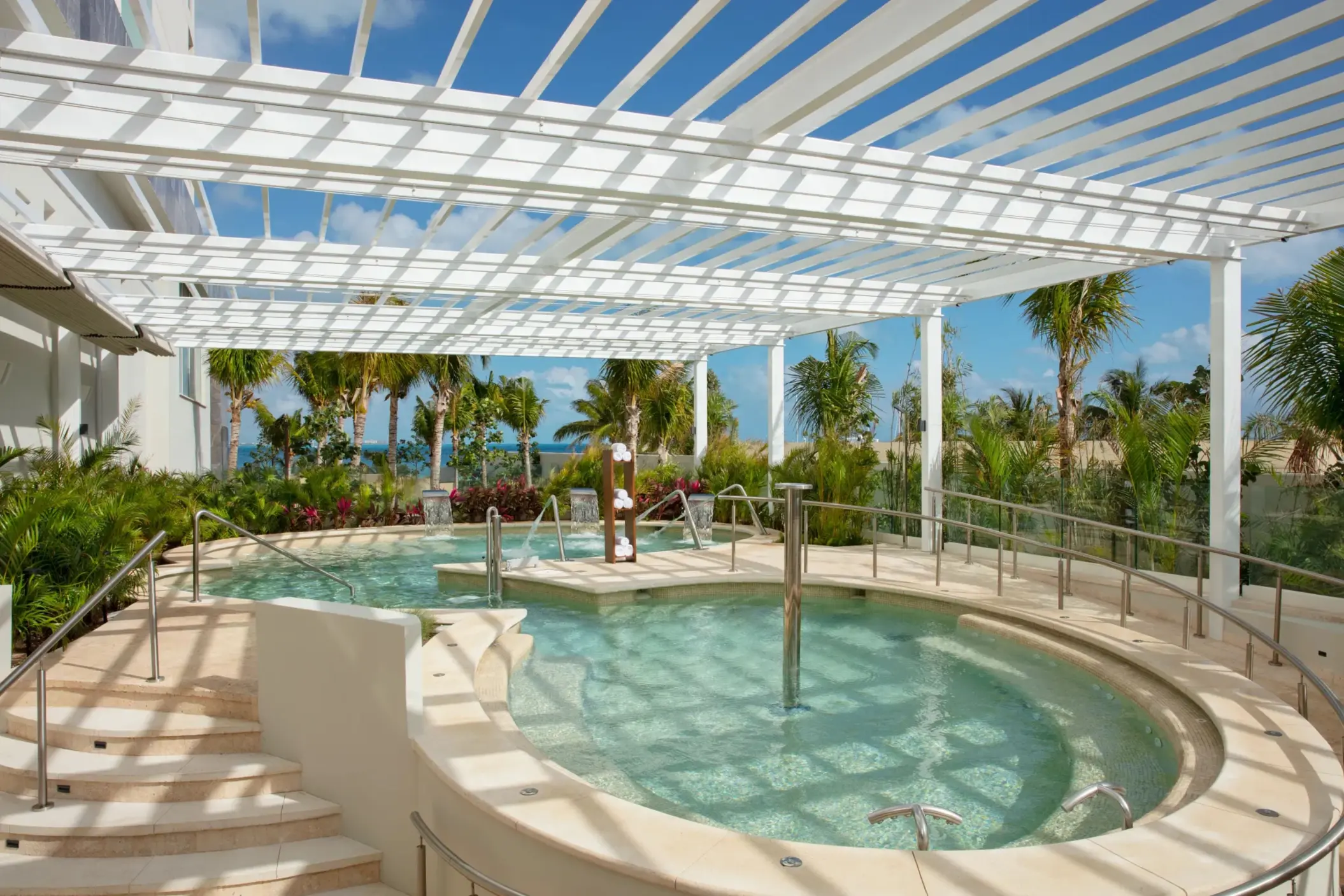 Outdoor hydrotherapy pool surrounded by palm trees and white pergola with ocean in the background.
