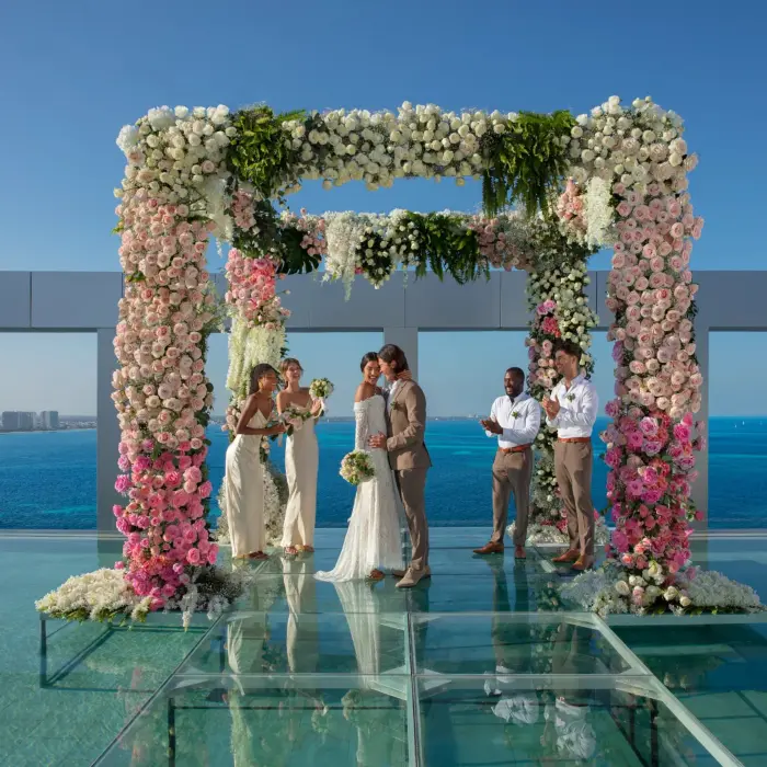 Bride and groom share a kiss under floral wedding arch on rooftop terrace overlooking turquoise ocean.