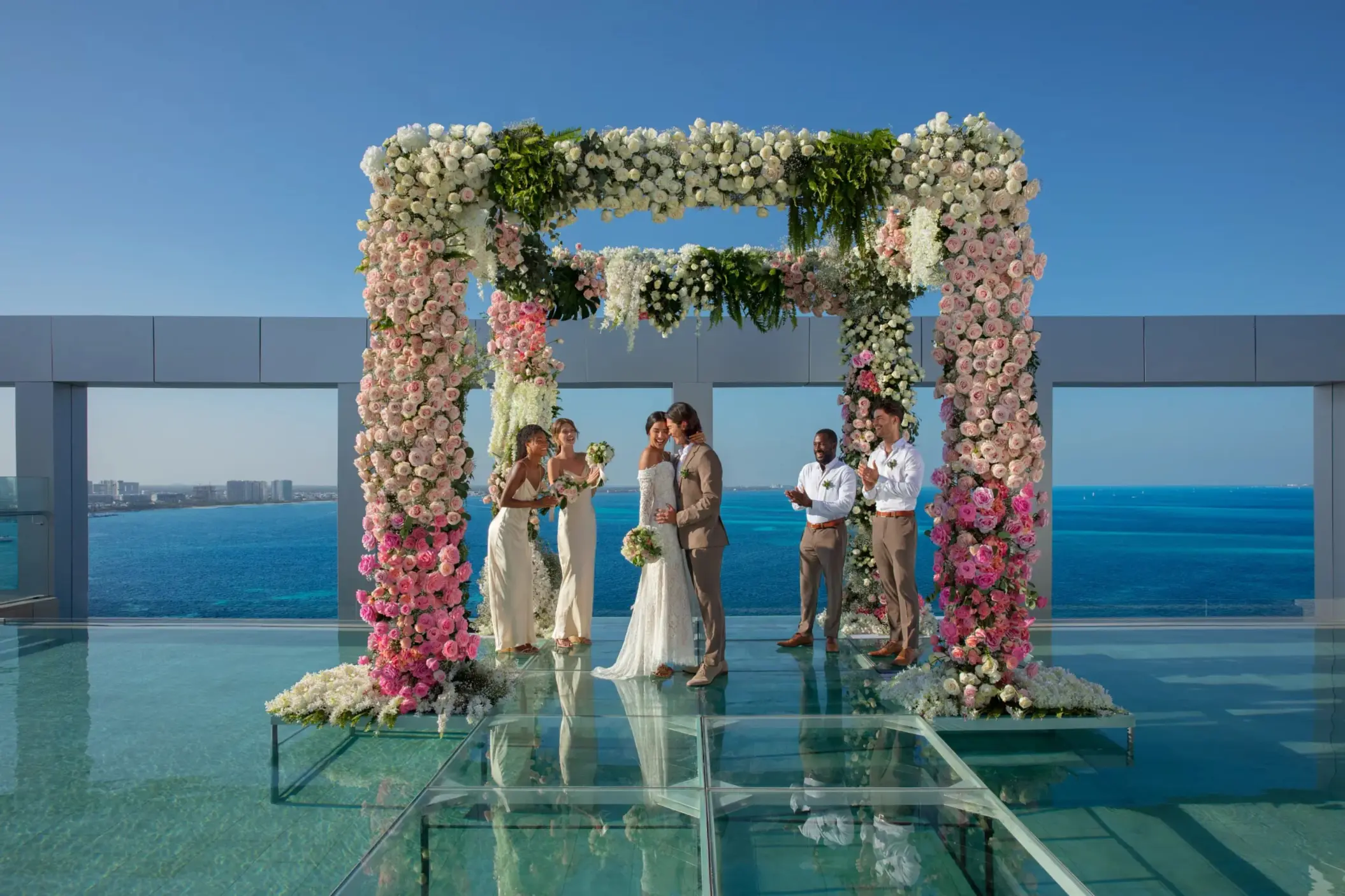 Bride and groom share a kiss under floral wedding arch on rooftop terrace overlooking turquoise ocean.