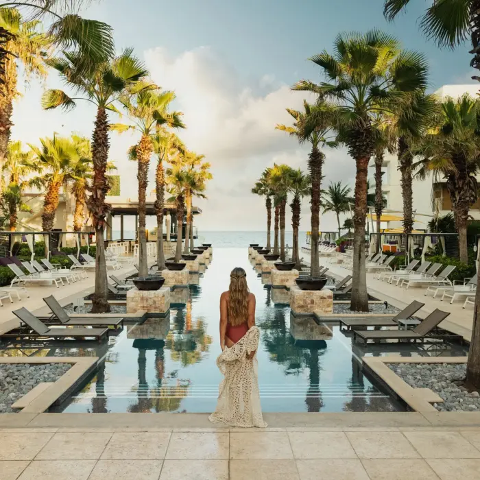 Woman at sunrise facing an infinity pool lined with palm trees and ocean backdrop.