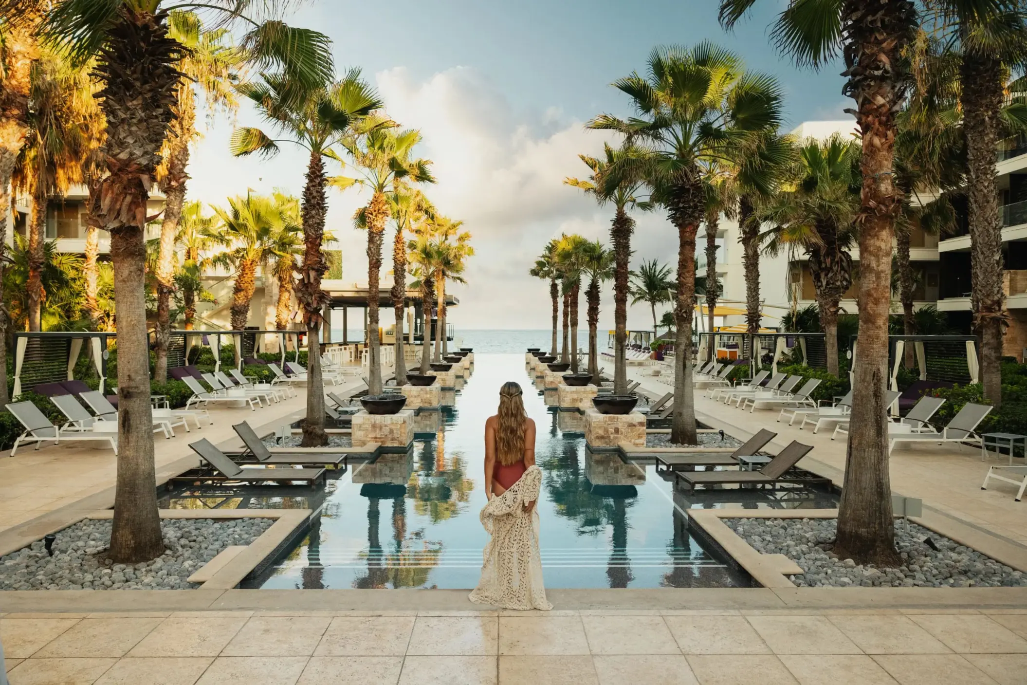 Woman at sunrise facing an infinity pool lined with palm trees and ocean backdrop.