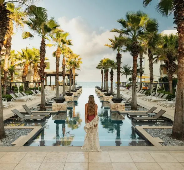 Woman at sunrise facing an infinity pool lined with palm trees and ocean backdrop.