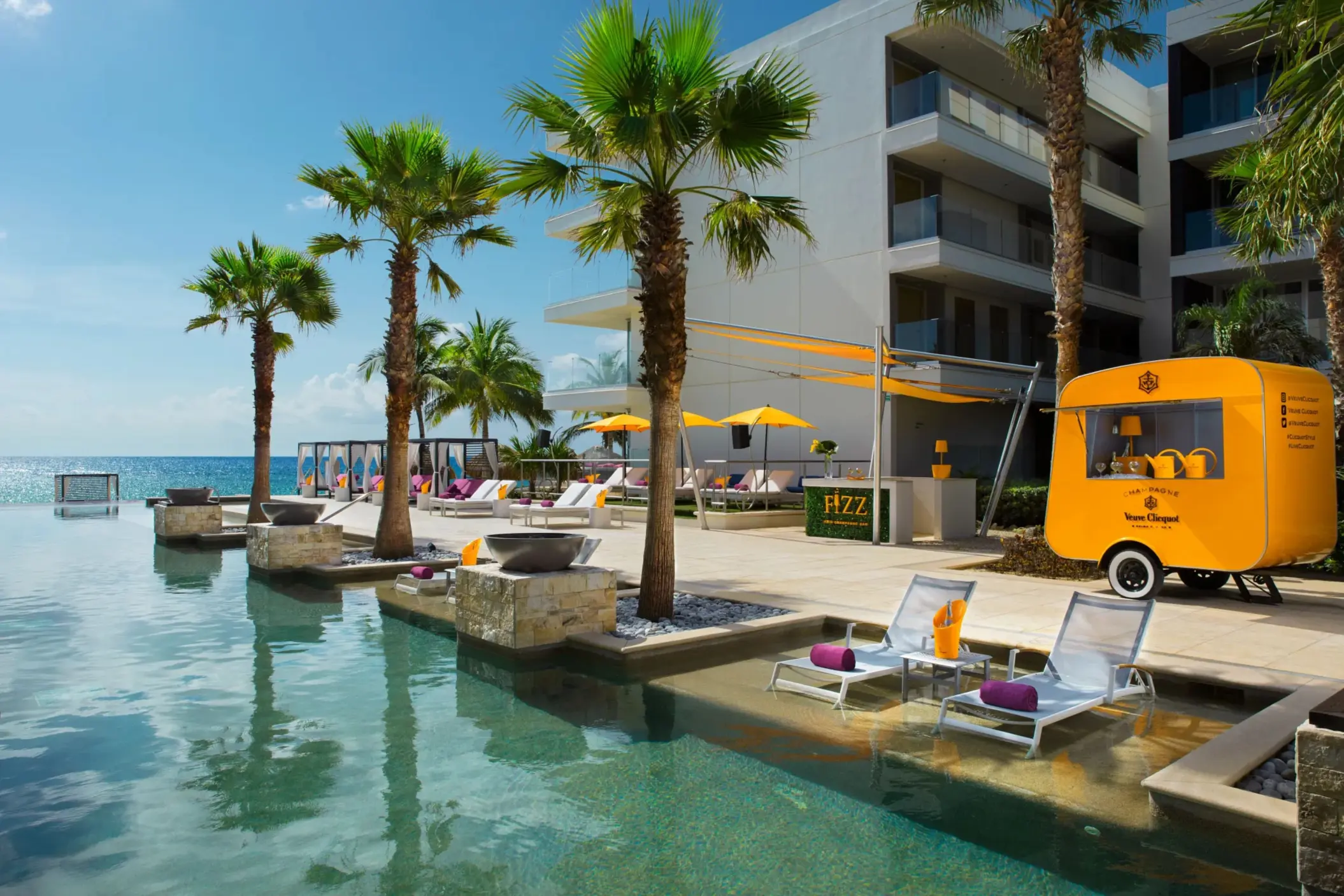 Bright yellow Veuve Clicquot cart and champagne bar beside sunny beachfront pool.