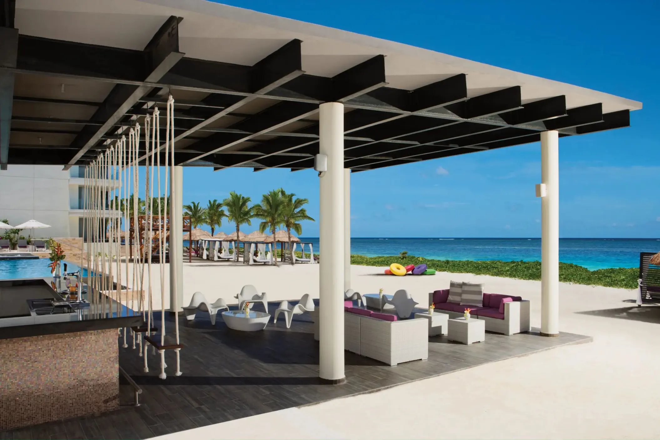 Swing-seat beach bar with lounge furniture overlooking white sand and turquoise ocean.