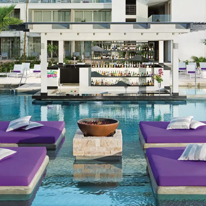 Colorful pool bar with floating purple loungers and views of the resort buildings.
