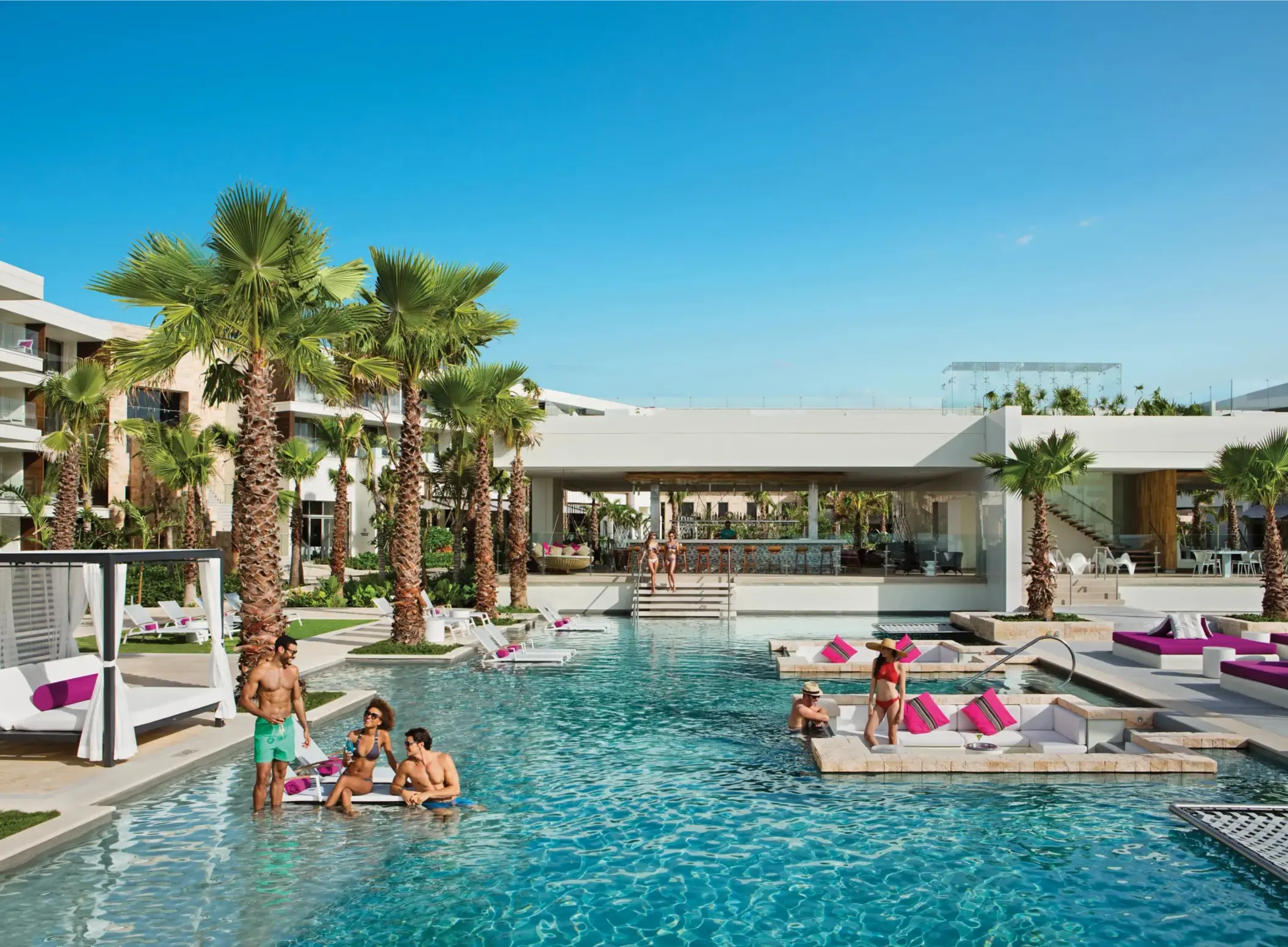 Guests socializing in the main pool near swim-up bar and cabanas under sunny skies.