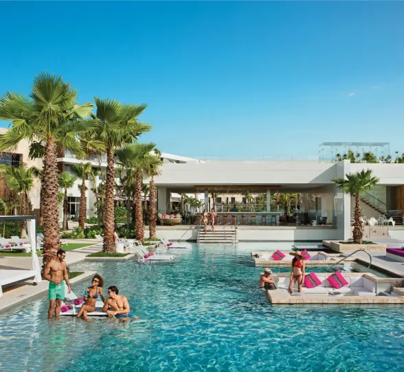 Guests socializing in the main pool near swim-up bar and cabanas under sunny skies.