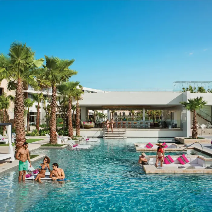 Guests socializing in the main pool near swim-up bar and cabanas under sunny skies.