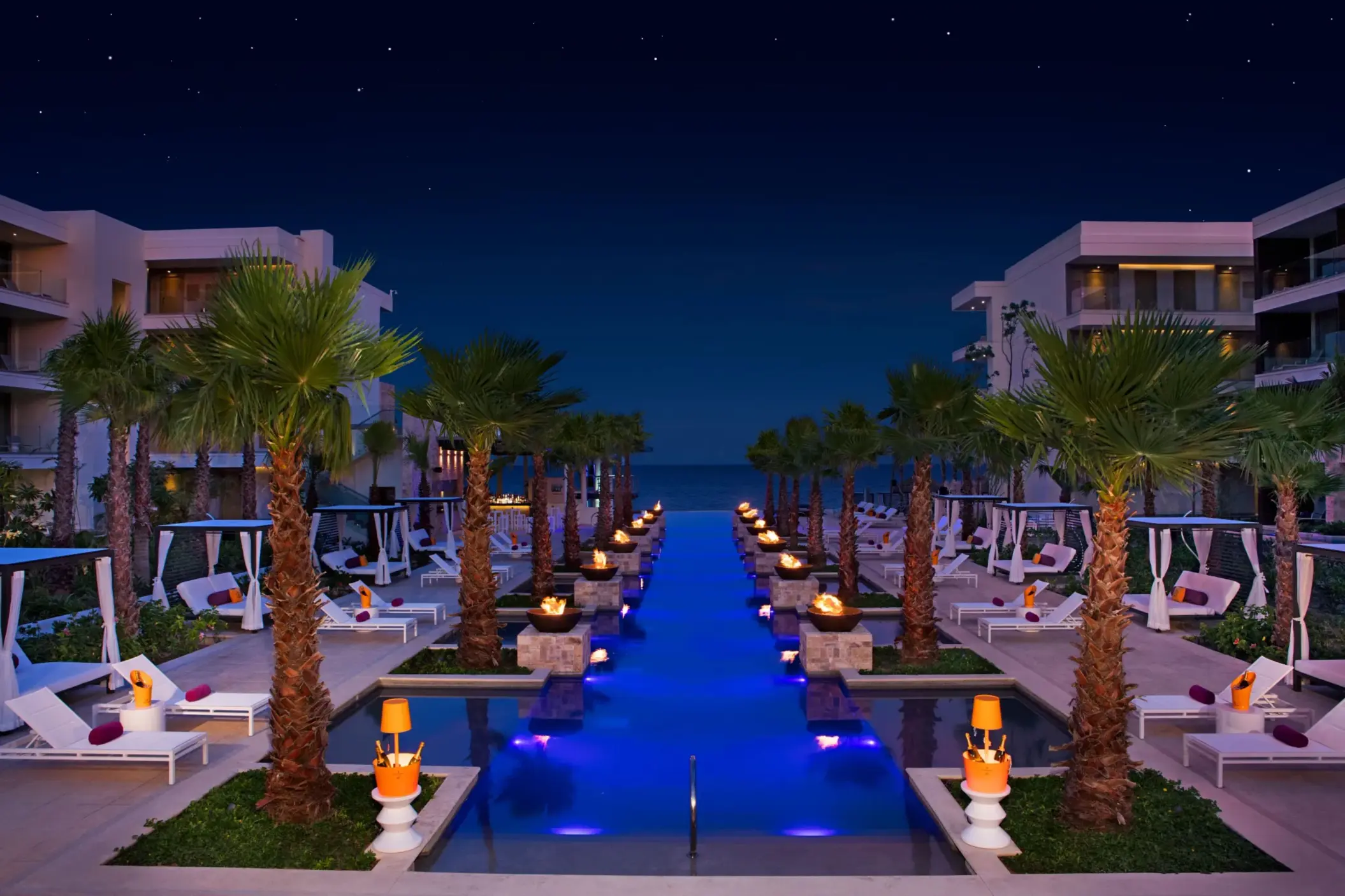 Fire-lit infinity pool scene at night with cabanas, palm trees, and ocean in the distance.