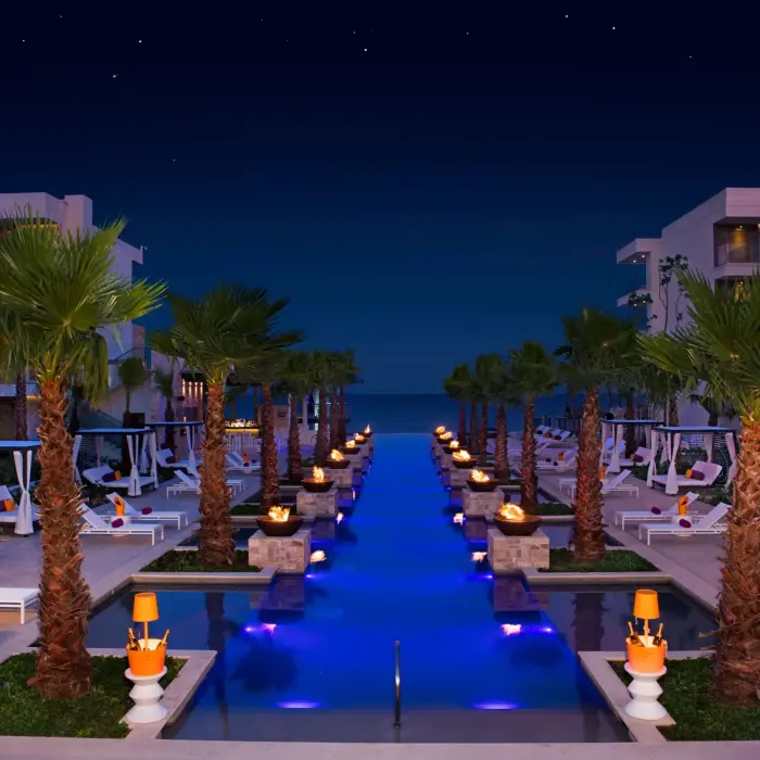Fire-lit infinity pool scene at night with cabanas, palm trees, and ocean in the distance.